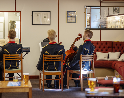 The Unwinding Room aboard Britannia. There are three mannequins, viewed from behind. They are sitting on chairs with instruments and music stands. In the foreground there are tables with drinks on it.
