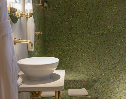 A green tiled bathroom aboard Fingal, featuring a shower, white round sink and brass fixtures. 