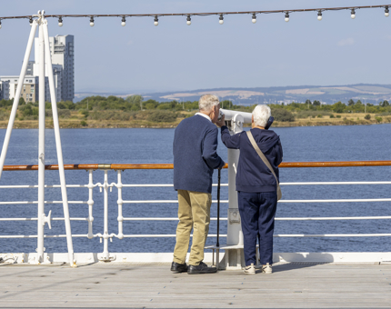 Two visitors are on Britannia's Verandah Deck looking through a telescope. There are views over the Port of Leith and the Firth of Forth. 