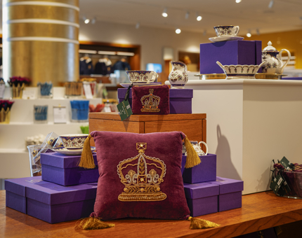 A look at some of the products in the new Shop. There is a maroon pillow with a gold embroidered crown on it. There is also a set of china. 