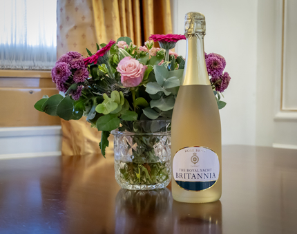 A bottle of Britannia pink fizz on a table next to a vase of flowers. 
