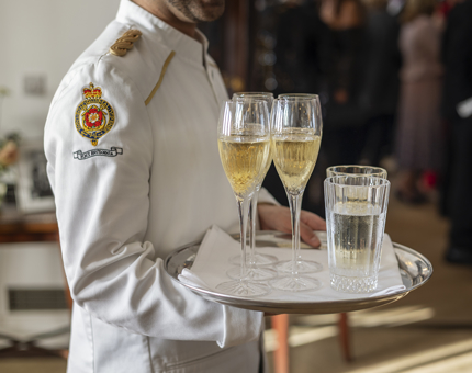 A server wearing a white coat with a Britannia crest on it is holding a tray with Champagne glasses and other drinks. 