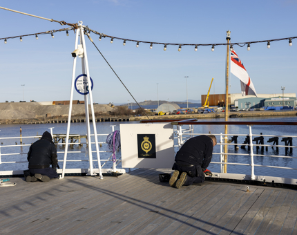 Two men kneeling on the Verandah Deck painting the railings white. You can see the Port of Leith in the background. 