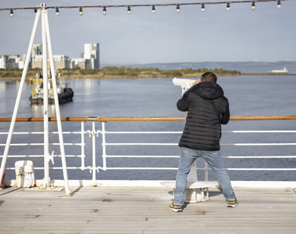 A man on the Verandah Deck looking through a telescope at views of the Port of Leith. 