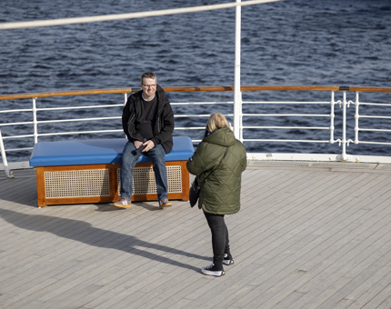 Two visitors on the Verandah Deck, one is sitting down, the other is standing, You can see the water in the background.