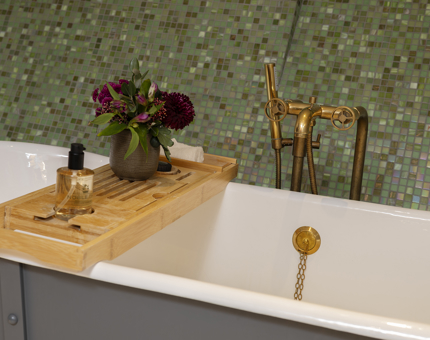 A bathtub with a wooden tray over it holding a vase of purple flowers. There are green tiles on the wall in the background. 