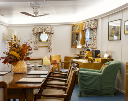 A warm, wood‑panelled room with nautical touches, featuring a large table with chairs, a desk with a lamp, a green armchair, and two porthole windows with patterned curtains. A vase of autumn‑coloured flowers sits on the table, adding to the cosy, lived‑in feel.
