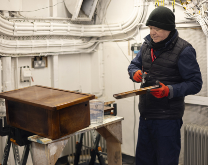 A maintenance man varnishing a wooden step from behind Britannia's Bell on the Verandah Deck. 
