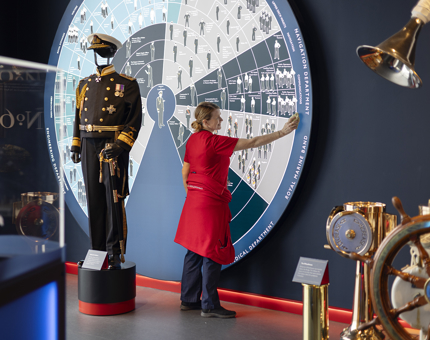 A Housekeeper is wiping a round information sign in The Royal Yacht Britannia's Visitor Centre. There is a mannequin in a Naval uniform displayed in front of the sign. 