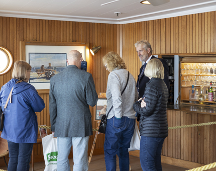 4 visitors are in the Sun Lounge talking to a Visitor Assistant aboard The Royal Yacht Britannia. 