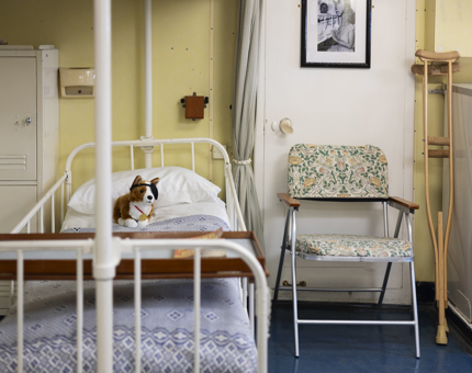 The bed in the onboard Sick Bay. There is a chair next to the bed and crutches. There is a corgi soft toy on the bed. 