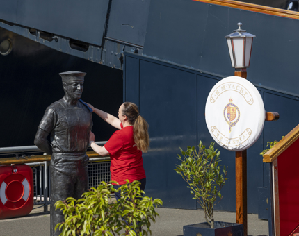 A Housekeeper is on the quayside next to Britannia cleaning a statue of a sailor. 