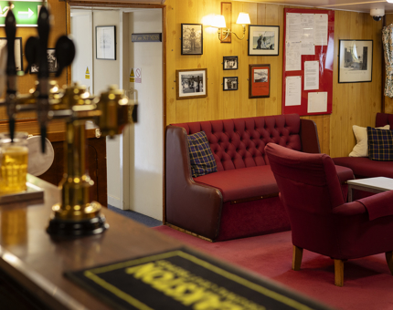 A view of an onboard Officers Mess. There is a bar, and the room is decorated in dark red furnishings. 