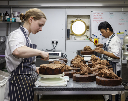 Two Chefs are in Britannia's Galley, they are icing chocolate cakes. 