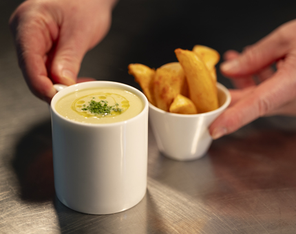 Hands placing a mug of soup and a bowl of potato wedges on a counter top.