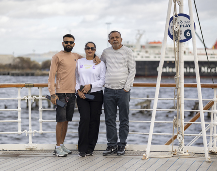 3 Visitors pose for a photo onboard Britannia in the Port of Leith. There is a ferry vessel in the background. 