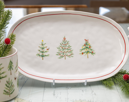An oval shaped plate with three Christmas tree illustrations on in. There is a small ceramic pot with that same illustrations on it and a plant in it too. 