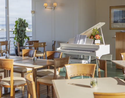 In the Royal Deck Tearoom there are tables and chairs set to welcome guests. There is a white grand piano in the background. 