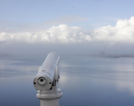 A foggy view over the water from the Verandah Deck. There is a white telescope in the foreground. The fog and clouds are very low and close to the water. 