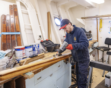 A Maintenance man is in the workshop using a heat gun to remove varnish off of a handrail. 