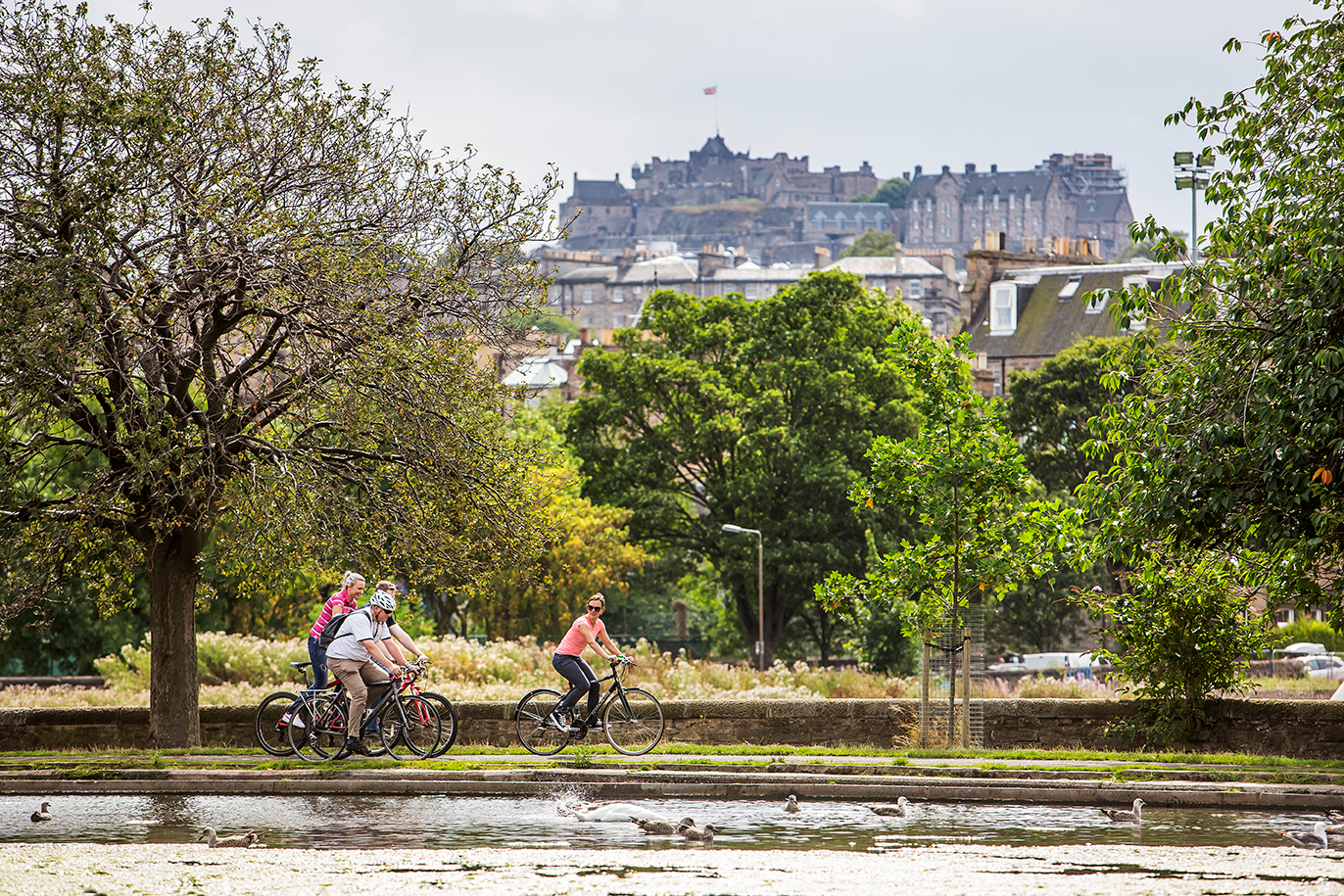 A group of cyclists are riding bikes in front of a pond a trees. Edinburgh Castle is in the background.