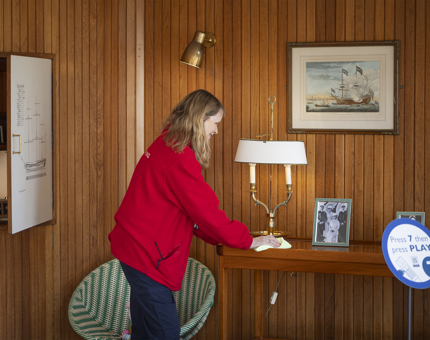 A Housekeeper wearing a red fleece is holding a cloth, wiping down a wooden table. She is in the Sun Lounge, it has wood paneled walls with a framed picture of a ship.