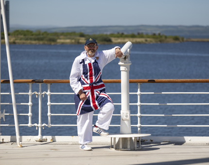 A Yottie taking a break on the Verandah Deck. He is wearing white overalls and a union jack apron. He is leaning on a telescope next to the railings. 