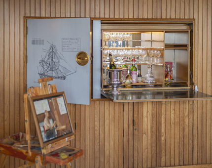 The drinks cabinet in the Sunlounge aboard The Royal Yacht Britannia. There is also an easel with a painting on it set up in the foreground. 