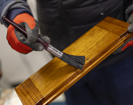 A close up of a hand holding a paintbrush, applying varnish to a wooden step. 