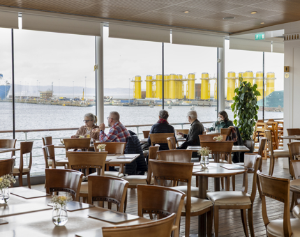 Guests are sitting at tables by the large windows in the Royal Deck Tearoom aboard Britannia. 