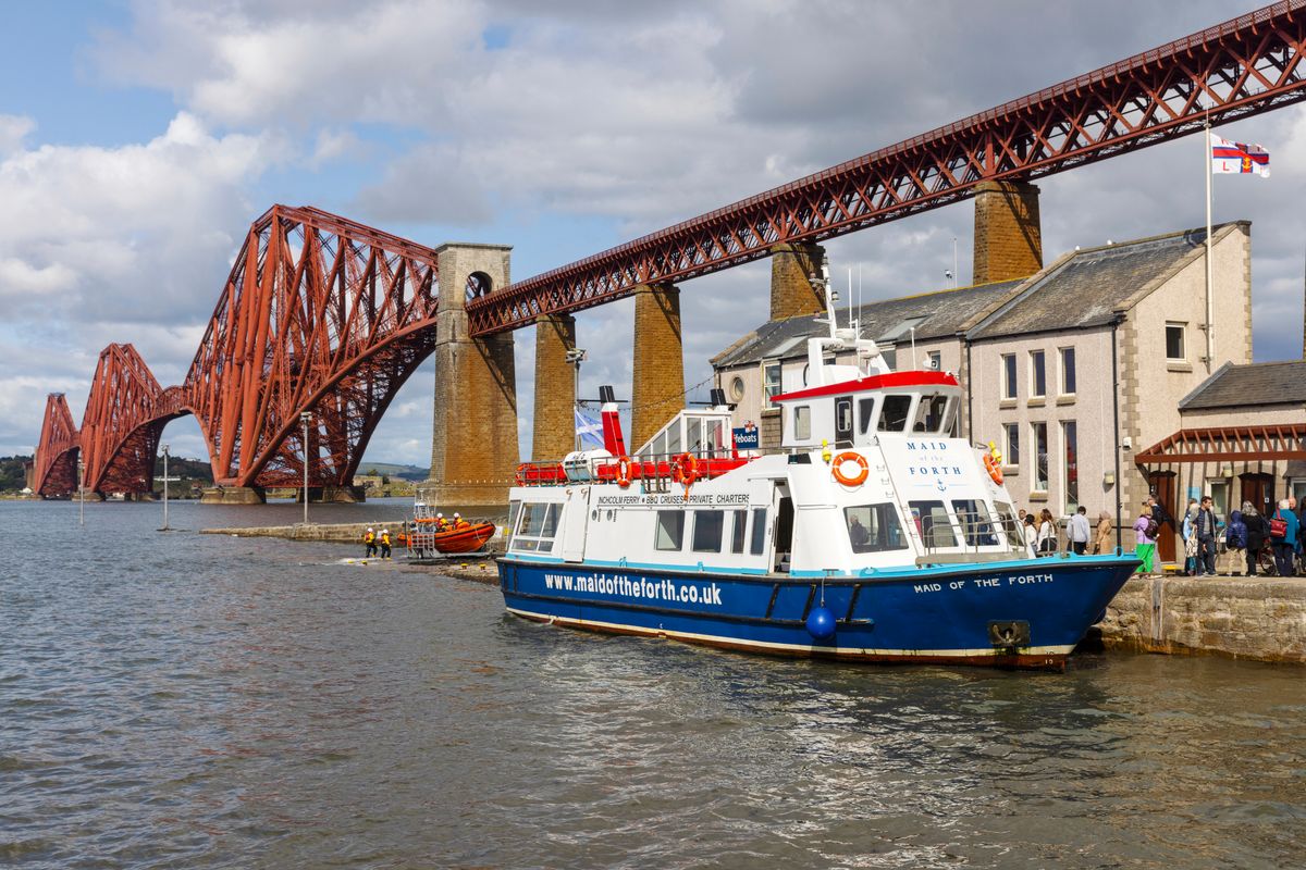 A Forth Boat Tours boat is docked with the Forth Road Bridge in the background on a sunny day in Edinburgh.