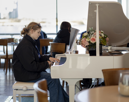 In the Royal Deck Tearoom a visitor is playing the white grand piano. There is a bunch of pink and white flowers sitting on the edge. 