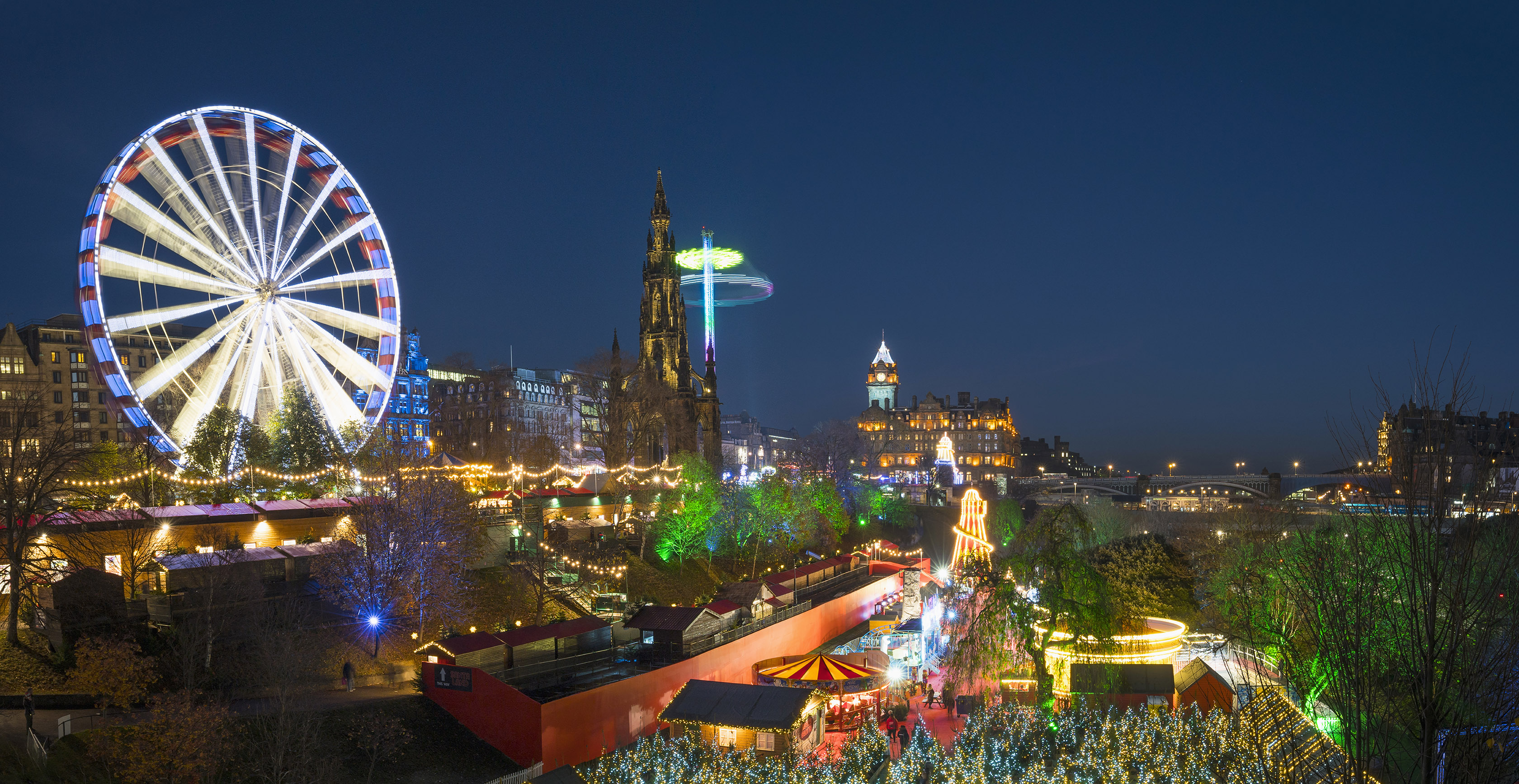 A view of Princes Street Gardens with Christmas Markets. It is night and there are colourful lights. There is a ferris wheel and Starflyer.