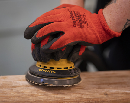 A close up of hands wearing red safety gloves sanding a wooden handrail. 