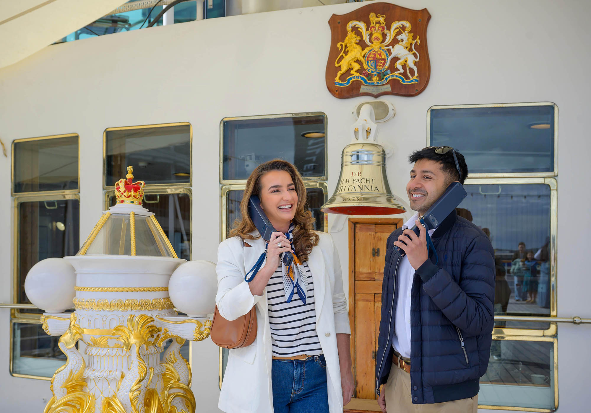 Two visitors stand on the deck beside the Britannia bell engraved “H.M. Yacht Britannia,” each holding a vintage-style telephone receiver. Behind them is a wooden plaque with the royal coat of arms and an ornate gold‑and‑white decorative structure topped with a crown.