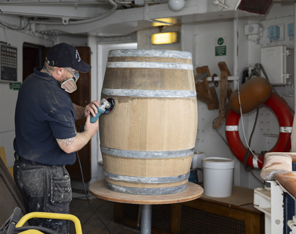 A wooden barrel is being sanded by a Maintenance tam member, He is holding an electric sander and wearing a facemask.