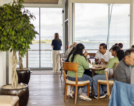 Visitors sitting at tables in the Royal Deck Tearoom. A visitor is standing by the floor to ceiling windows in the background, looking at the view of the Port of Leith, Edinburgh. 