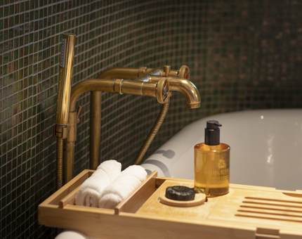 A close up of a bath, with a wooden bath tray that has small white towels and a Noble Isle bath product on it. 