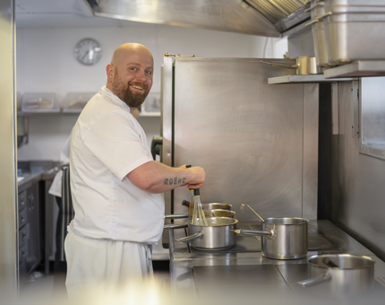 A Chef is standing by a cooker, whisking a pot on the stove top. 