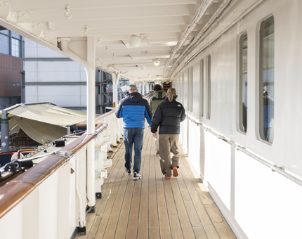 Three visitors walking on the deck of Britannia listening to audio guide handsets. 